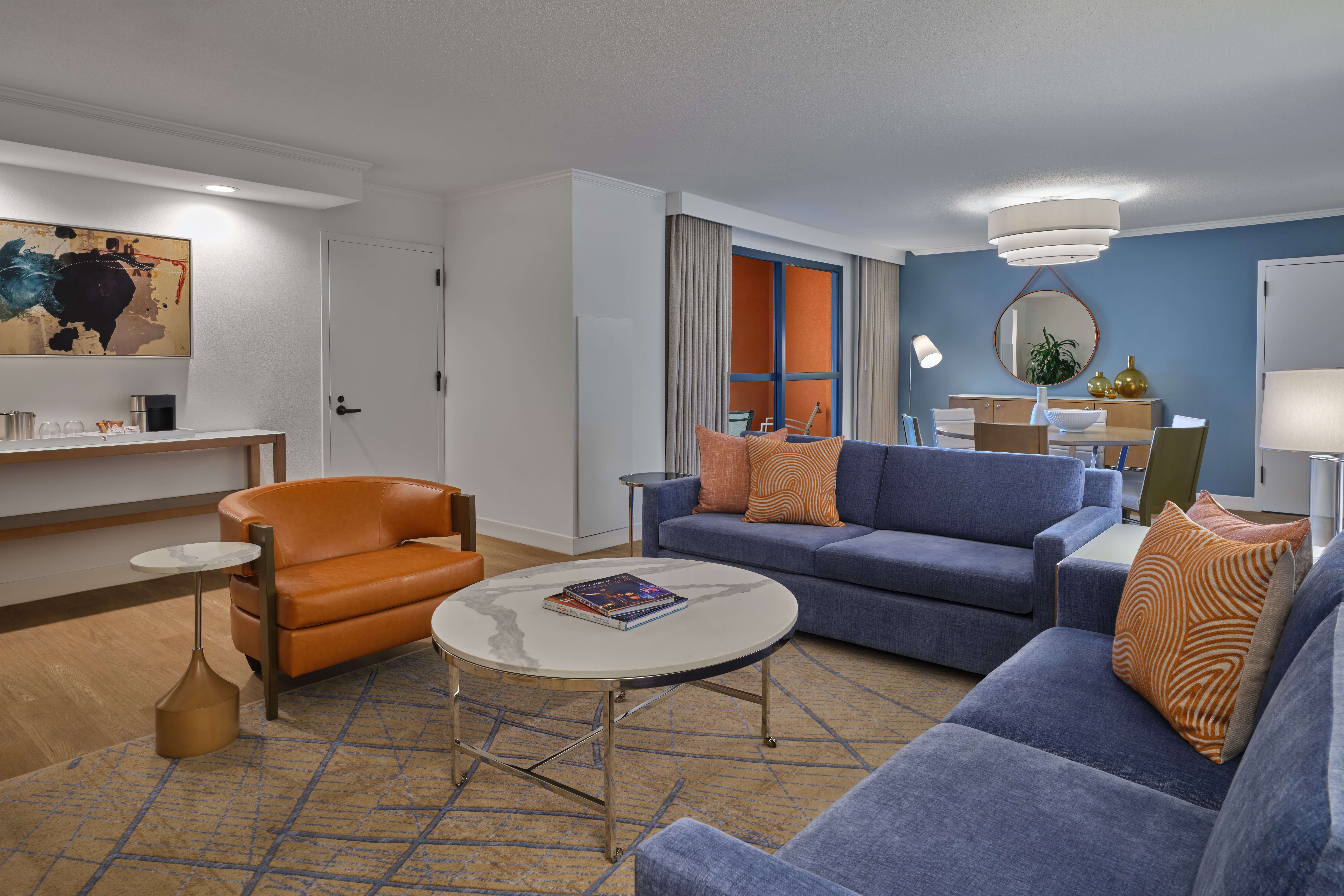 Modern living room with blue sofas, orange armchair, round marble coffee table, patterned rug, and wall art. In the background, a dining area with a round mirror and blue accent wall is visible.