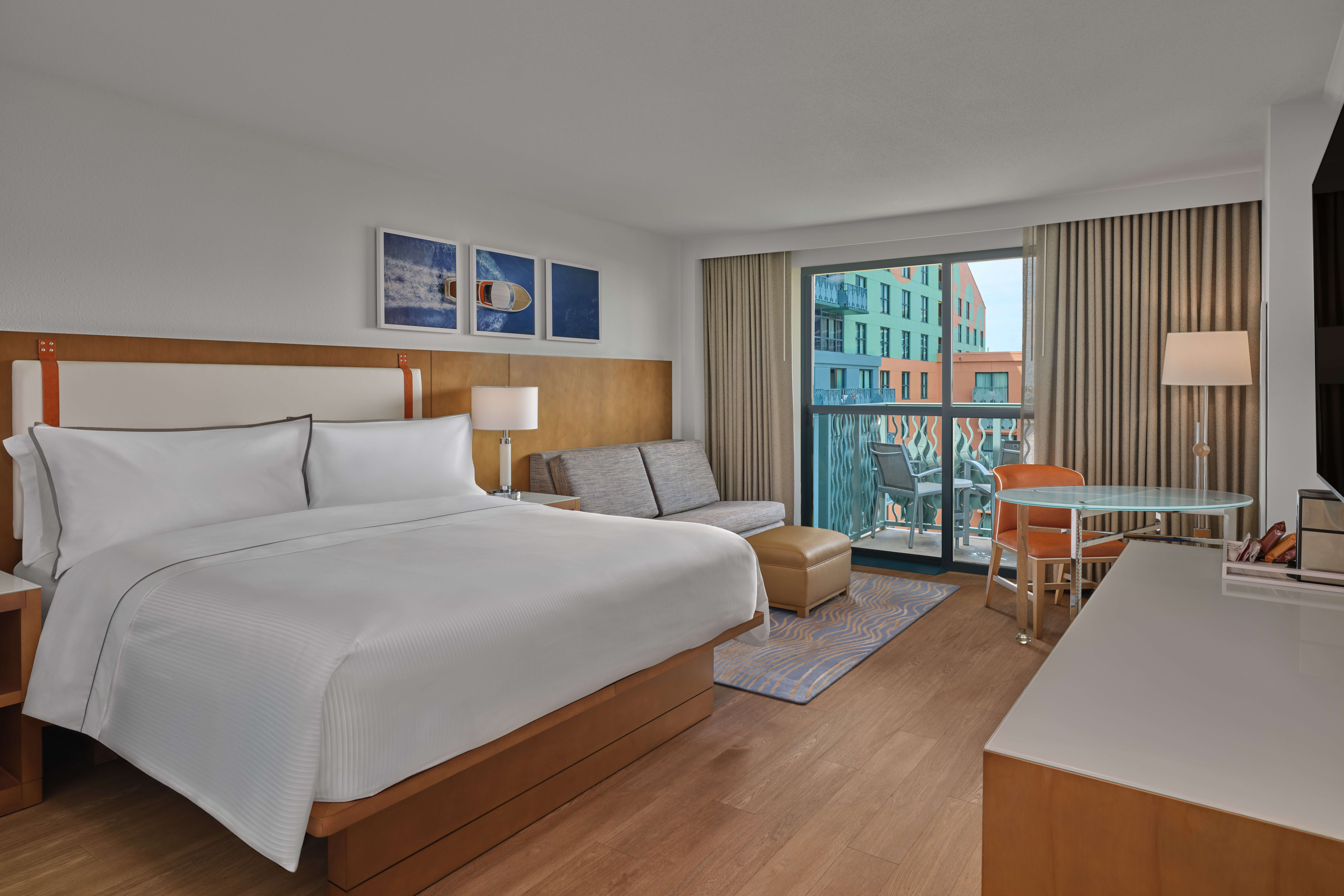 Modern hotel room with a large bed, sofa, ottoman, desk, and chair. Large windows with curtains reveal a balcony and colorful buildings outside. The room has wood floors and is decorated in neutral tones.