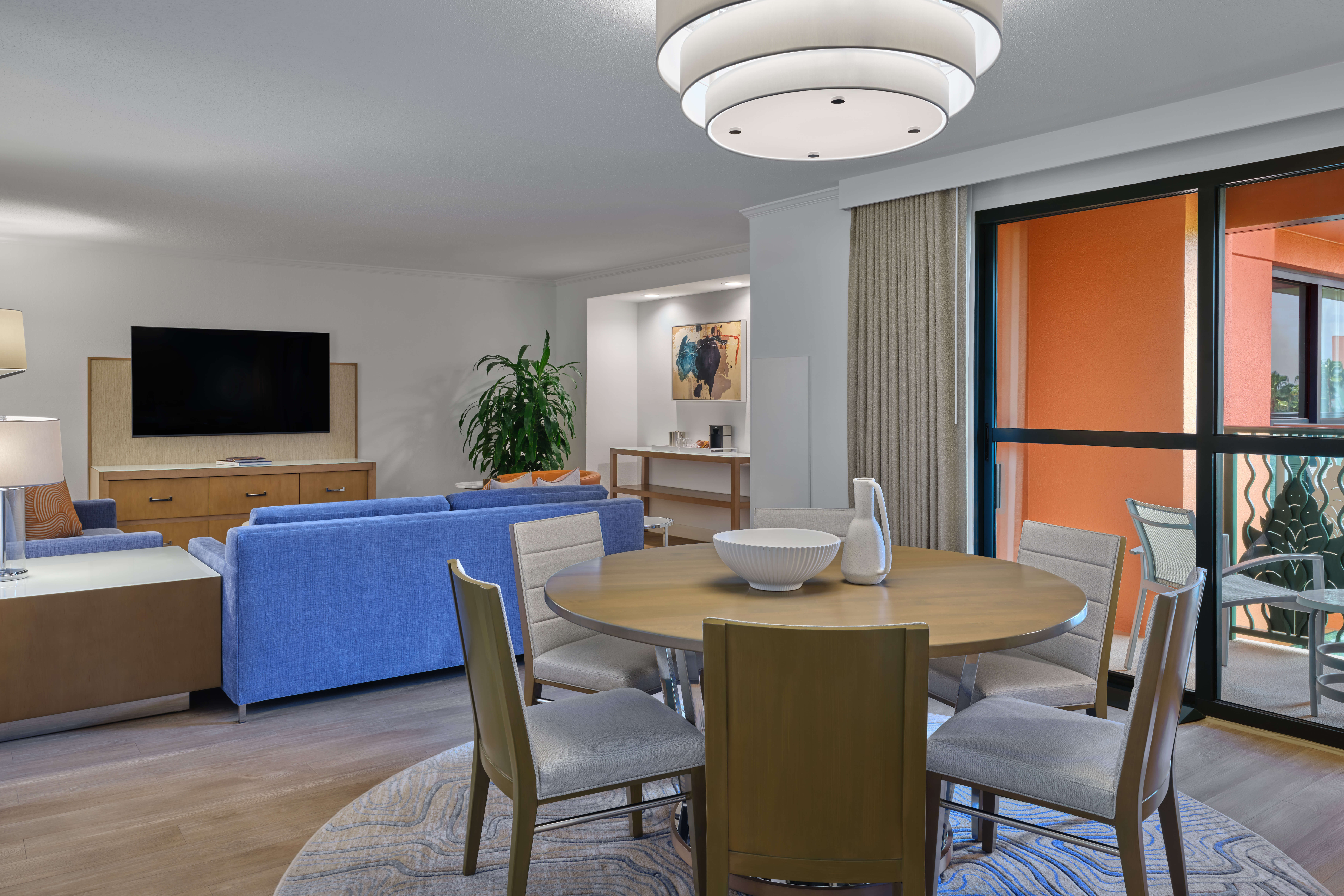 A modern living and dining area features a round wooden table with four chairs, a blue sofa facing a wall-mounted TV, a large potted plant, and sliding doors opening to an orange balcony. A chandelier hangs above the table.