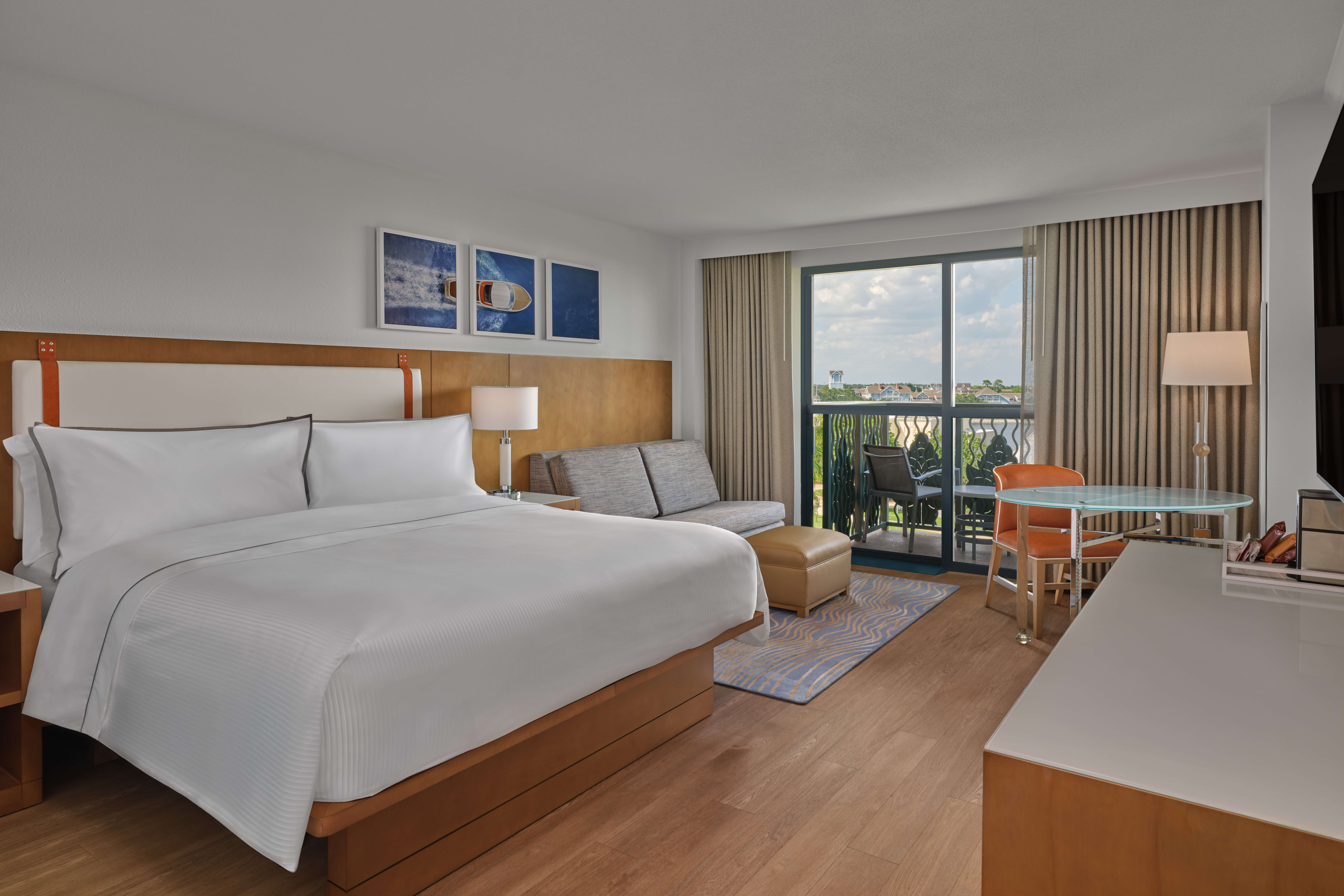 Modern hotel room with a large bed, a grey sofa, small table with chairs, desk, and a balcony with outdoor seating. Natural light comes through large windows with beige curtains. Artwork hangs above the bed.