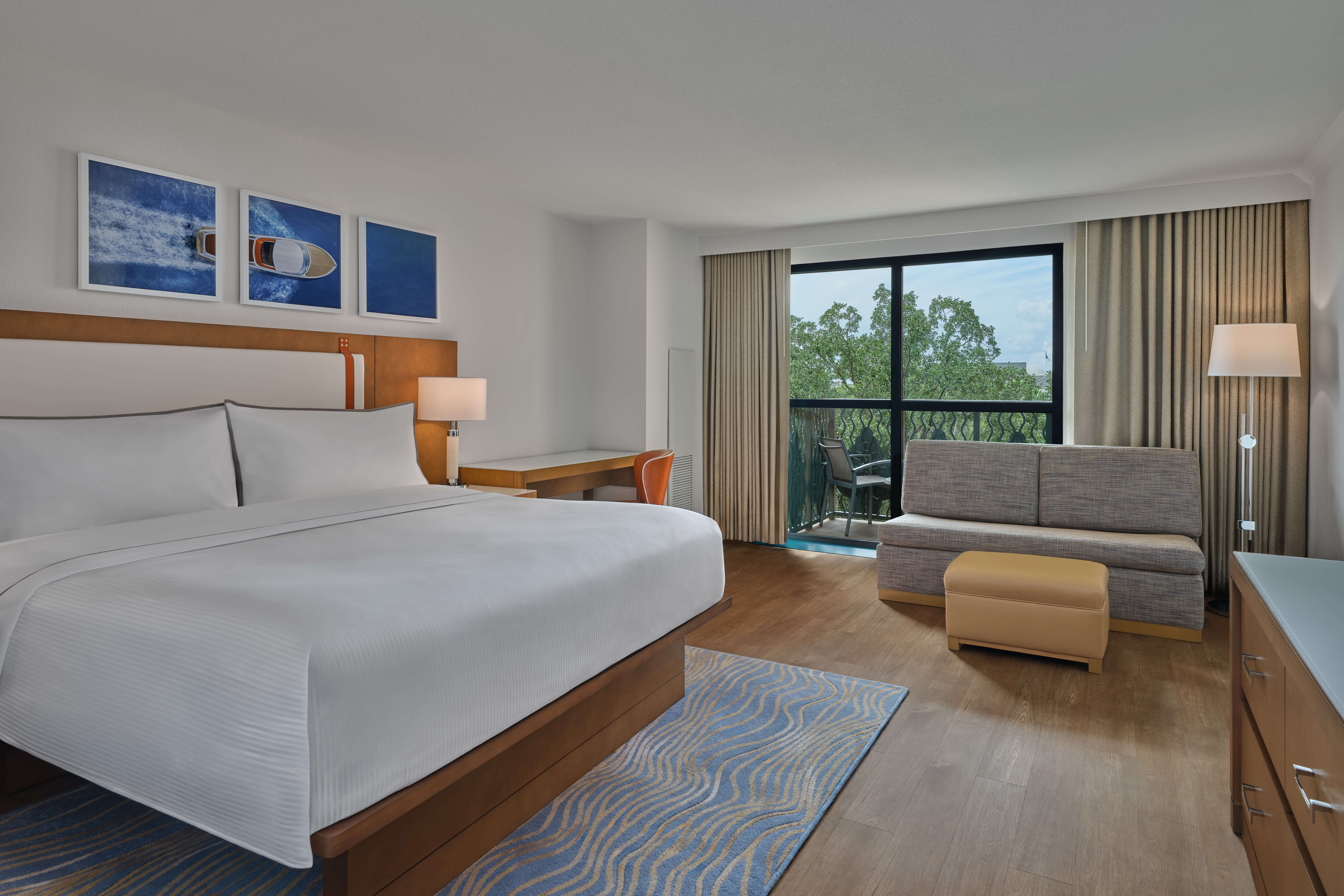 Modern hotel room with a large bed, desk, sofa, ottoman, floor lamps, and a balcony with glass doors overlooking trees. Light wood floors and blue-themed decor create a bright, airy atmosphere.