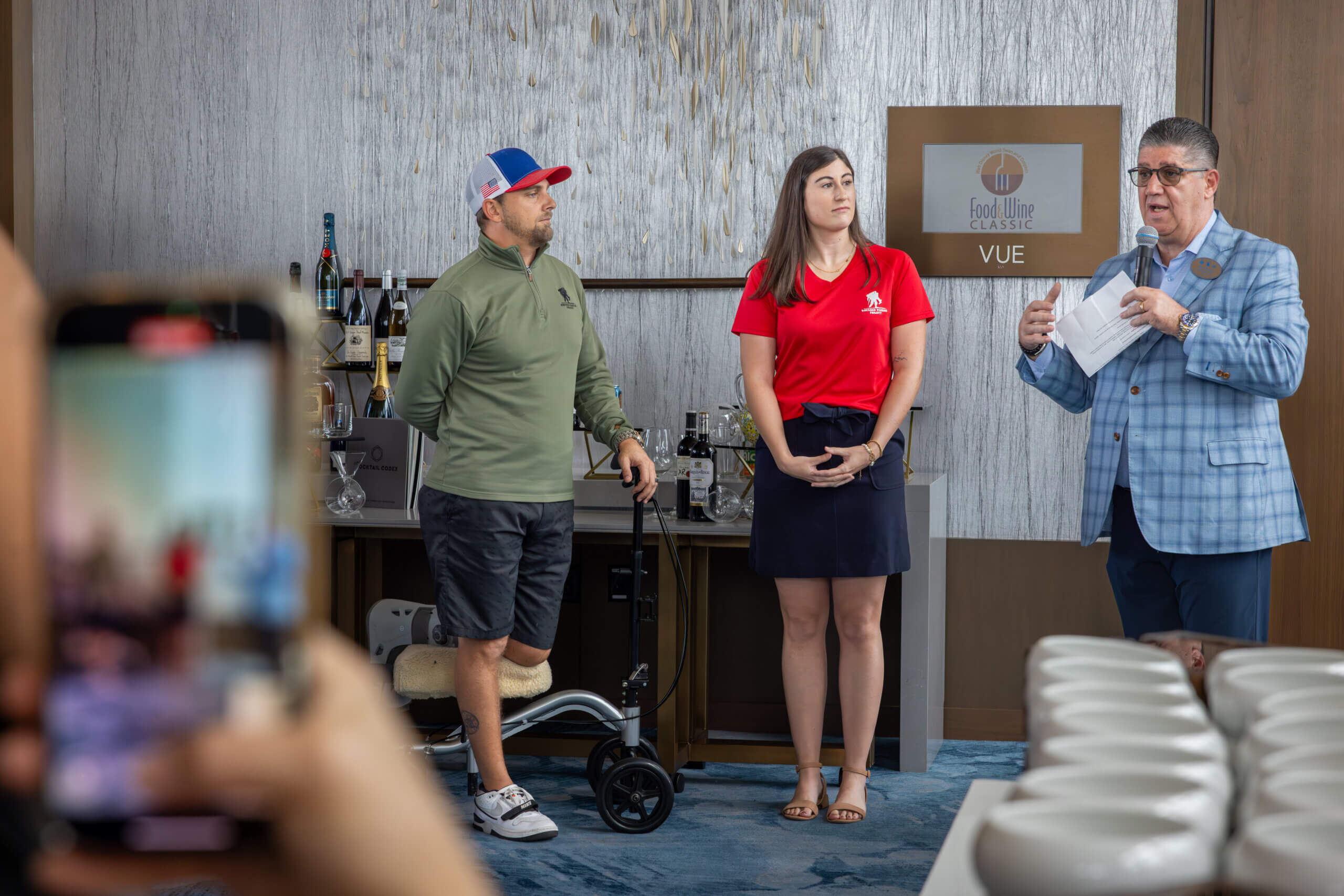 A man on a knee scooter, a woman in a red shirt, and a man in a blue blazer speak at an indoor event; bottles are on a table behind them and a person holds up a phone in the foreground.