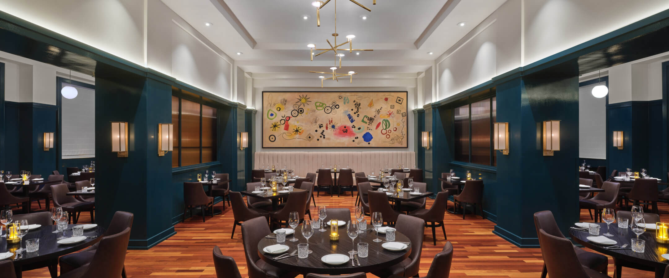 Upscale restaurant interior with dark wood floors, teal walls, modern chandeliers, and neatly set round tables. A large, colorful abstract artwork hangs on the far wall, creating a stylish and inviting atmosphere.