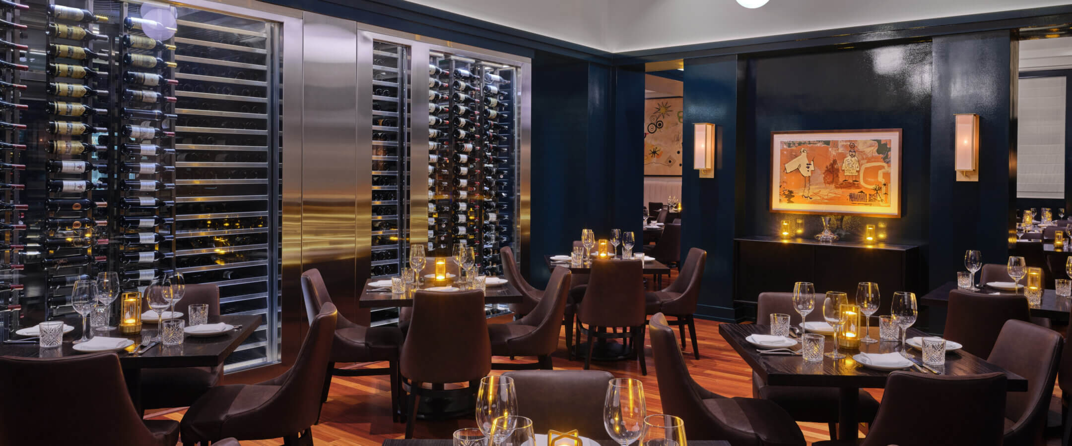 Elegant restaurant interior with dark wooden floors, brown leather chairs, set tables with glassware, and soft candle lighting. A large wall wine display and modern art add to the upscale ambiance.