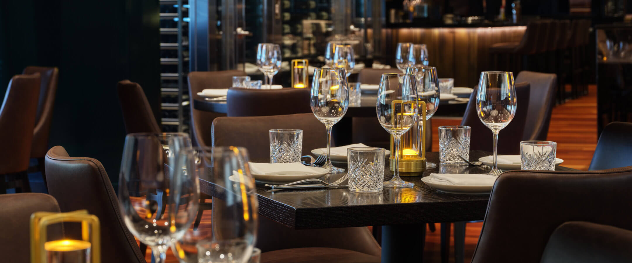 A modern, dimly lit restaurant with neatly set tables featuring wine glasses, plates, and candles. Leather chairs surround the tables, and the ambiance appears elegant and inviting.