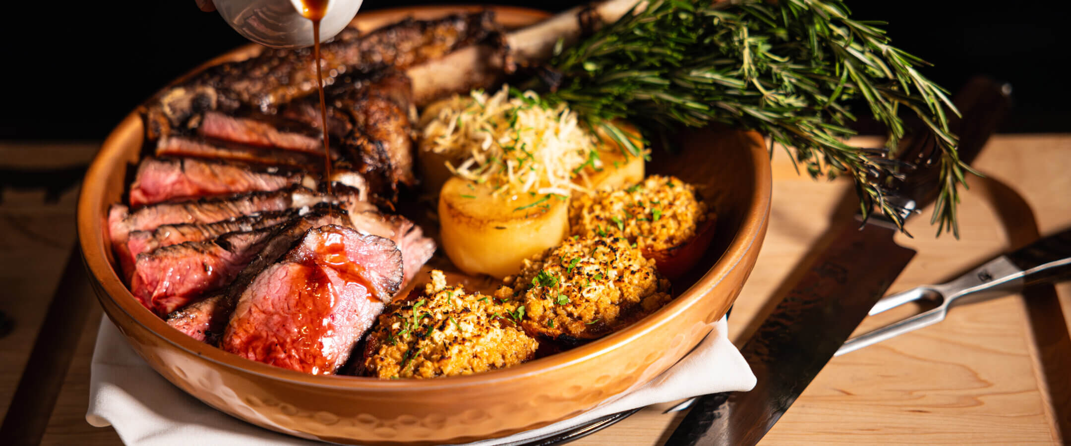 A hand pours sauce over sliced steak with a bone, served with herb potatoes, stuffed vegetables, and greens on a round plate. Knife and fork beside the dish. BOURBON STEAK is engraved on the wooden table.