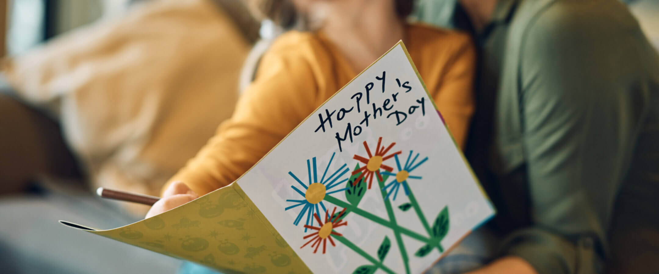 A child hugs and kisses a smiling woman, likely her mother, while holding a colorful handmade card that says Happy Mothers Day with drawings of flowers.