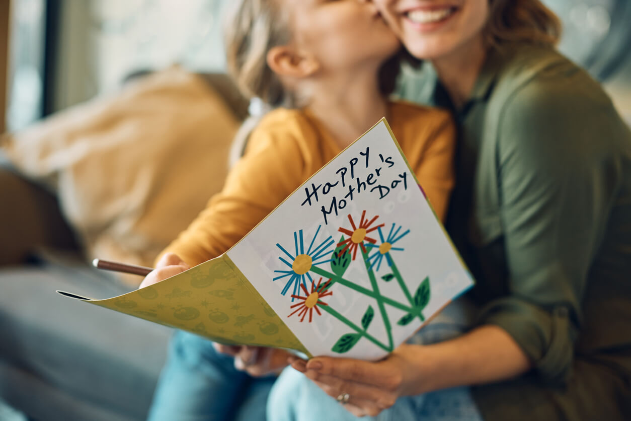 A child hugs and kisses a smiling woman, likely her mother, while holding a colorful handmade card that says Happy Mothers Day with drawings of flowers.