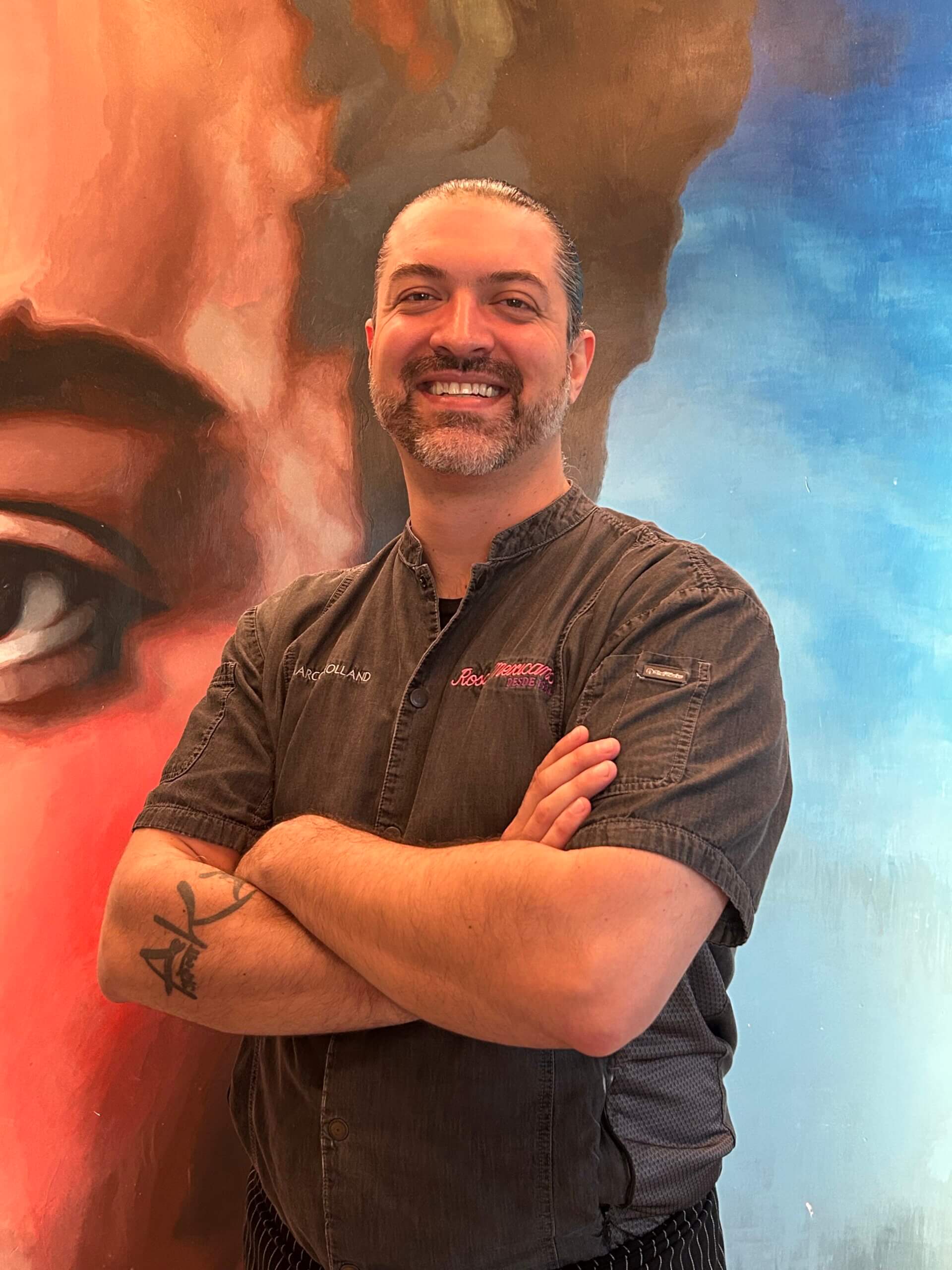 Ein lächelnder Mann mit Bart und Schnurrbart steht mit verschränkten Armen da und trägt eine dunkle Kochjacke. Im Hintergrund ist ein farbenfrohes Wandgemälde zu sehen, das ein Gesicht teilweise zeigt.