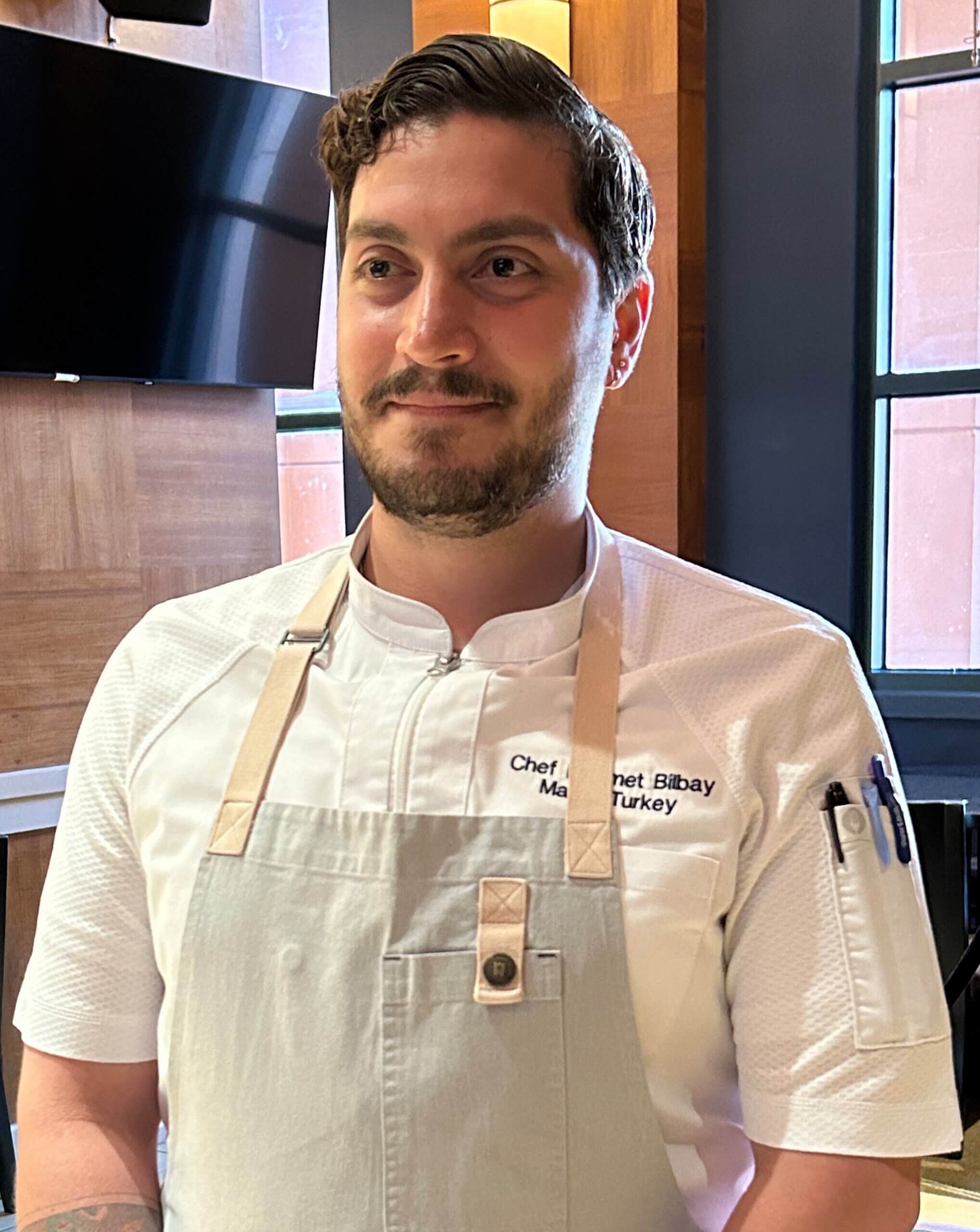 El chef Mehmet, vestido con una bata blanca y un delantal gris claro, se encuentra en el interior de un restaurante con paredes revestidas de madera, mirando ligeramente hacia un lado y sonriendo con dulzura.