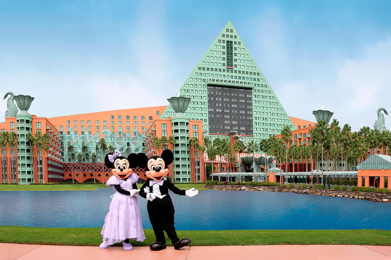 Mickey und Minnie Mouse stehen in festlicher Kleidung vor einem großen orange-grünen Hotel mit pyramidenförmigem Dach, umgeben von Palmen und einem Teich unter blauem Himmel.