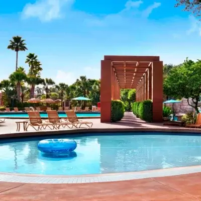 منطقة حمام سباحة في الهواء الطلق تضم lounge ومظلات خضراء وأشجار نخيل وحلقة زرقاء قابلة للنفخ تطفو في بركة صغيرة؛ ويؤدي ممر مقنطر من الطين إلى مسار محاط بالخضرة تحت سماء زرقاء صافية.