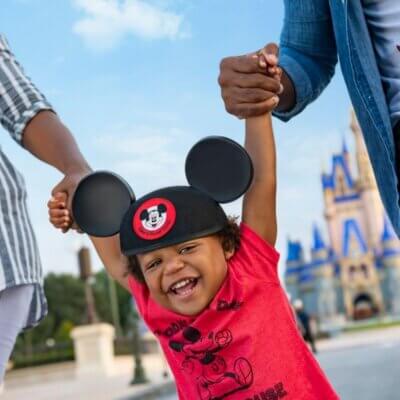 يظهر في الصورة طفل مبتسم يرتدي أذني ميكي ماوس وقميصًا أحمر، ويحمله شخصان بالذراعين أمام قلعة زرقاء، على الأرجح في إحدى مدن ملاهي ديزني.