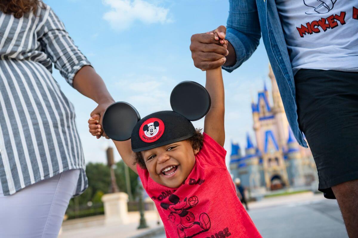 Ein lächelndes Kind mit Mickey-Mouse-Ohren und einem roten T-Shirt wird von zwei Erwachsenen vor einem blauen Schloss hochgehoben, vermutlich in einem Disney-Freizeitpark.