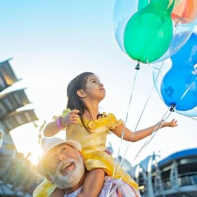 تجلس فتاة صغيرة ترتدي فستانًا أصفر على أكتاف رجل مسن، وهي تبتسم وتمسك بالونات ملونة في حفل يقام في الهواء الطلق، مع أشعة الشمس الساطعة والمباني في الخلفية.