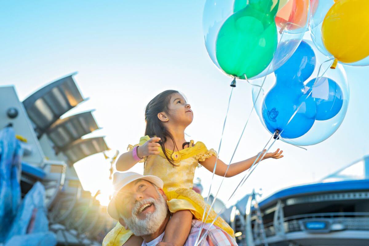 Ein junges Mädchen in einem gelben Kleid sitzt auf den Schultern eines älteren Mannes, lächelt und hält bunte Luftballons in der Hand; im Hintergrund sind strahlender Sonnenschein und Gebäude zu sehen.