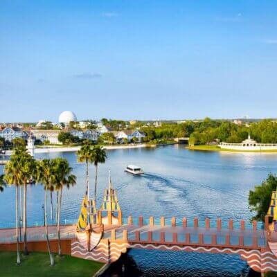 منظر خلاب لواجهة بحرية تضم جسراً مزخرفاً وأشجار نخيل وقارباً يبحر على الماء، مع مبانٍ ذات أسقف ملونة في الخلفية تحت سماء زرقاء صافية.