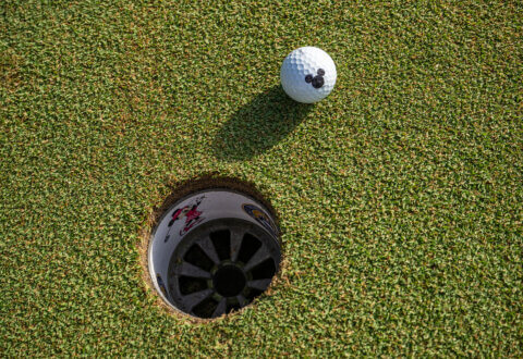 A golf ball with a Mickey Mouse logo rests near the edge of a hole on a putting green, with cartoon characters decorating the inside of the hole.