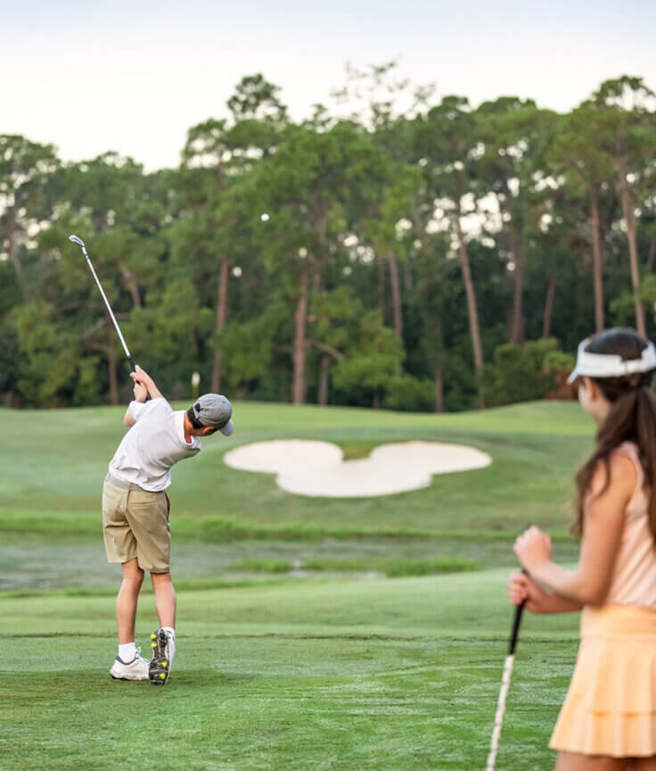 Un ragazzino sta facendo oscillare una mazza da golf su un campo rigoglioso, mentre tre persone, tra cui una ragazzina e due adulti, lo guardano giocare. Sullo sfondo si vedono degli alberi e un bunker di sabbia.