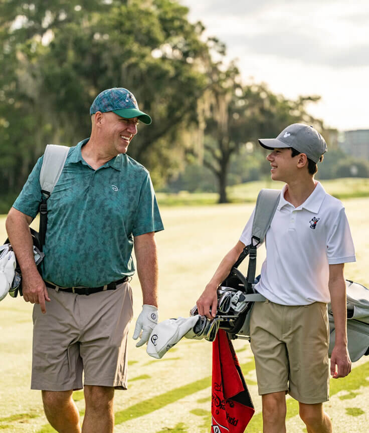 Due golfisti, un adulto e un adolescente, camminano fianco a fianco su un campo da golf inondato dal sole, con le loro sacche in mano e sorridendosi l'un l'altro. Sullo sfondo si intravedono alberi, una fontana e un edificio.