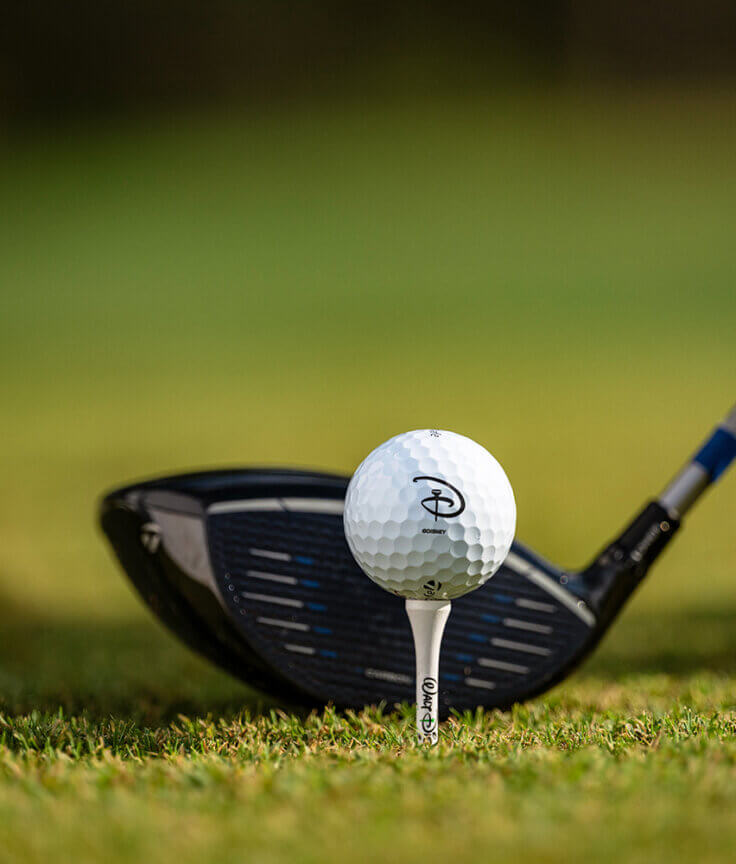 Primo piano di una mazza da golf e di una pallina da golf su un tee bianco, posizionate sull'erba verde, pronte per il primo colpo in una giornata soleggiata.
