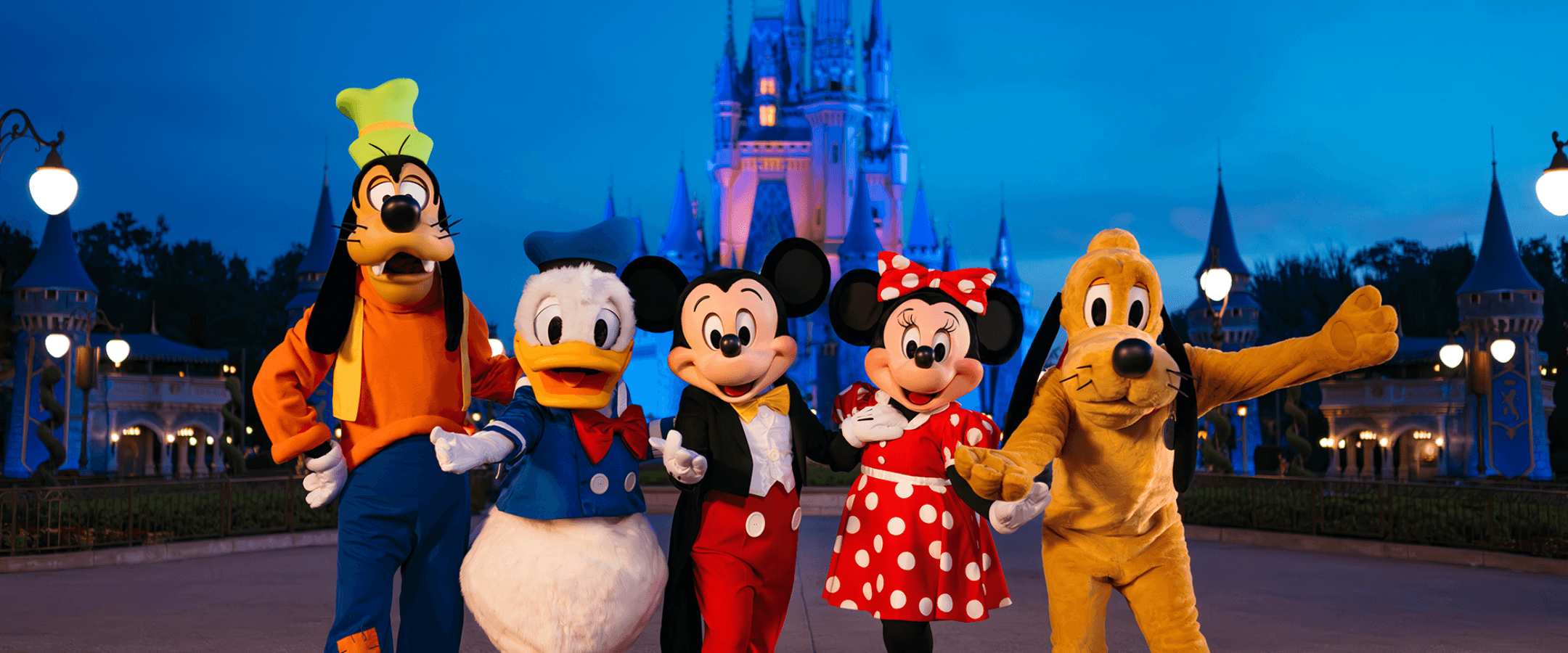 Five Disney characters—Goofy, Donald Duck, Mickey Mouse, Minnie Mouse, and Pluto—stand smiling in front of a brightly lit Cinderella Castle at dusk in a theme park.