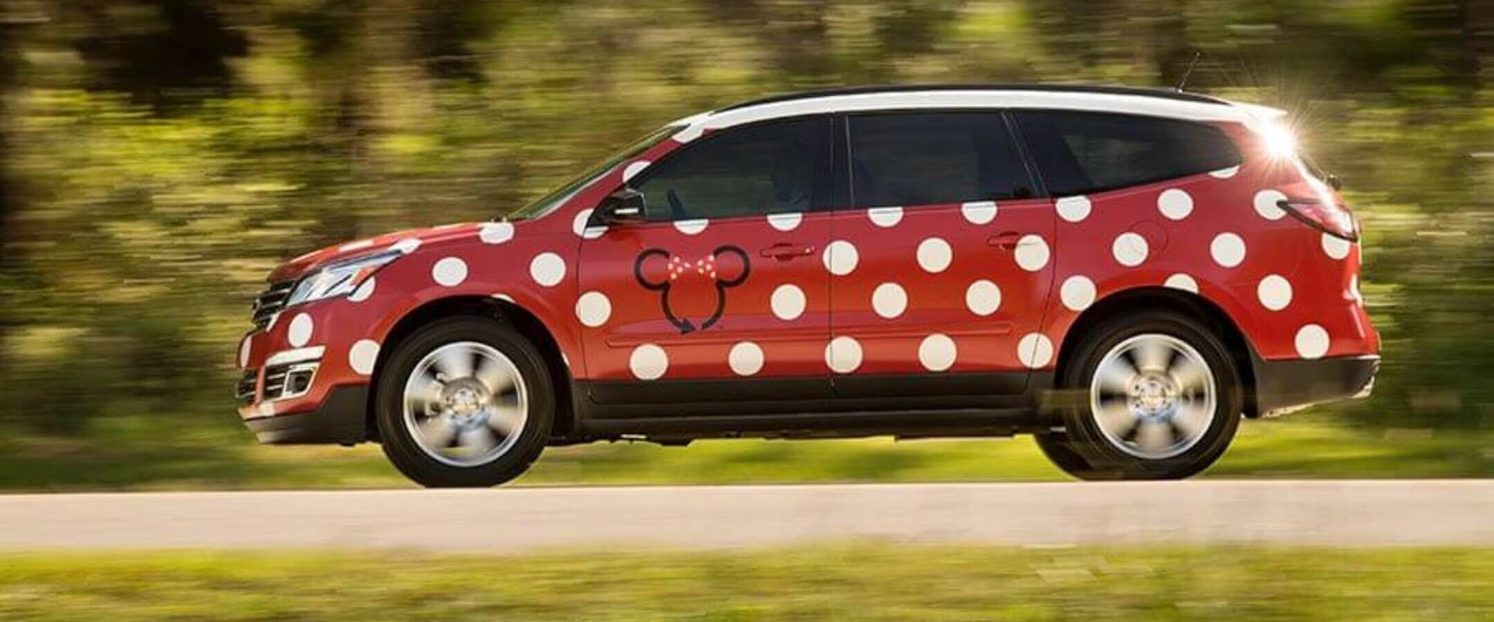 A red SUV with large white polka dots and a black outline of a bow on the side drives down a road with green trees blurred in the background.