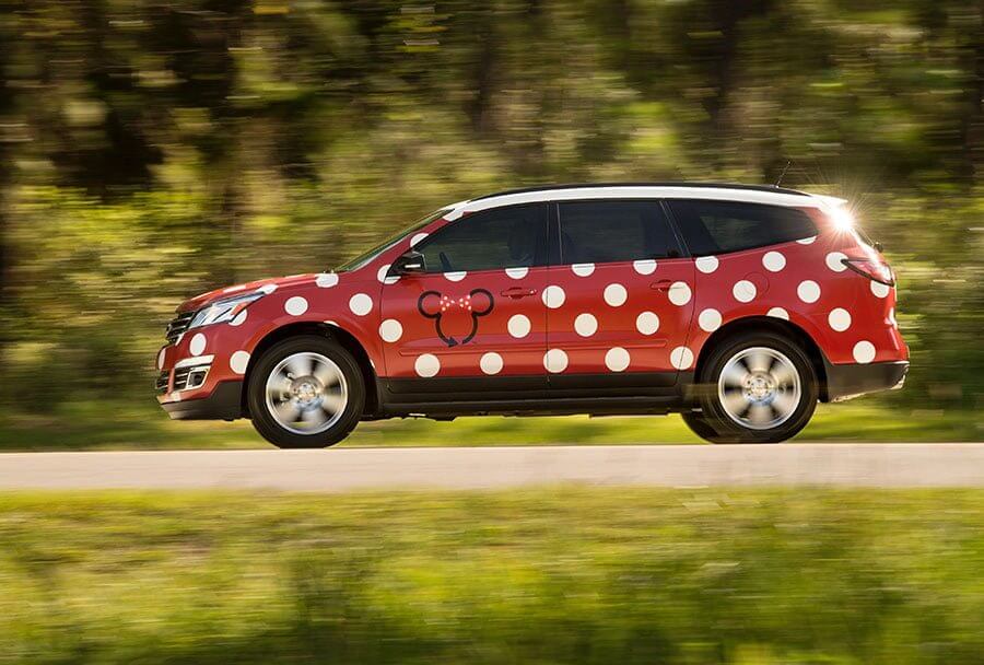 A red SUV with large white polka dots and a black outline of a bow on the side drives down a road with green trees blurred in the background.