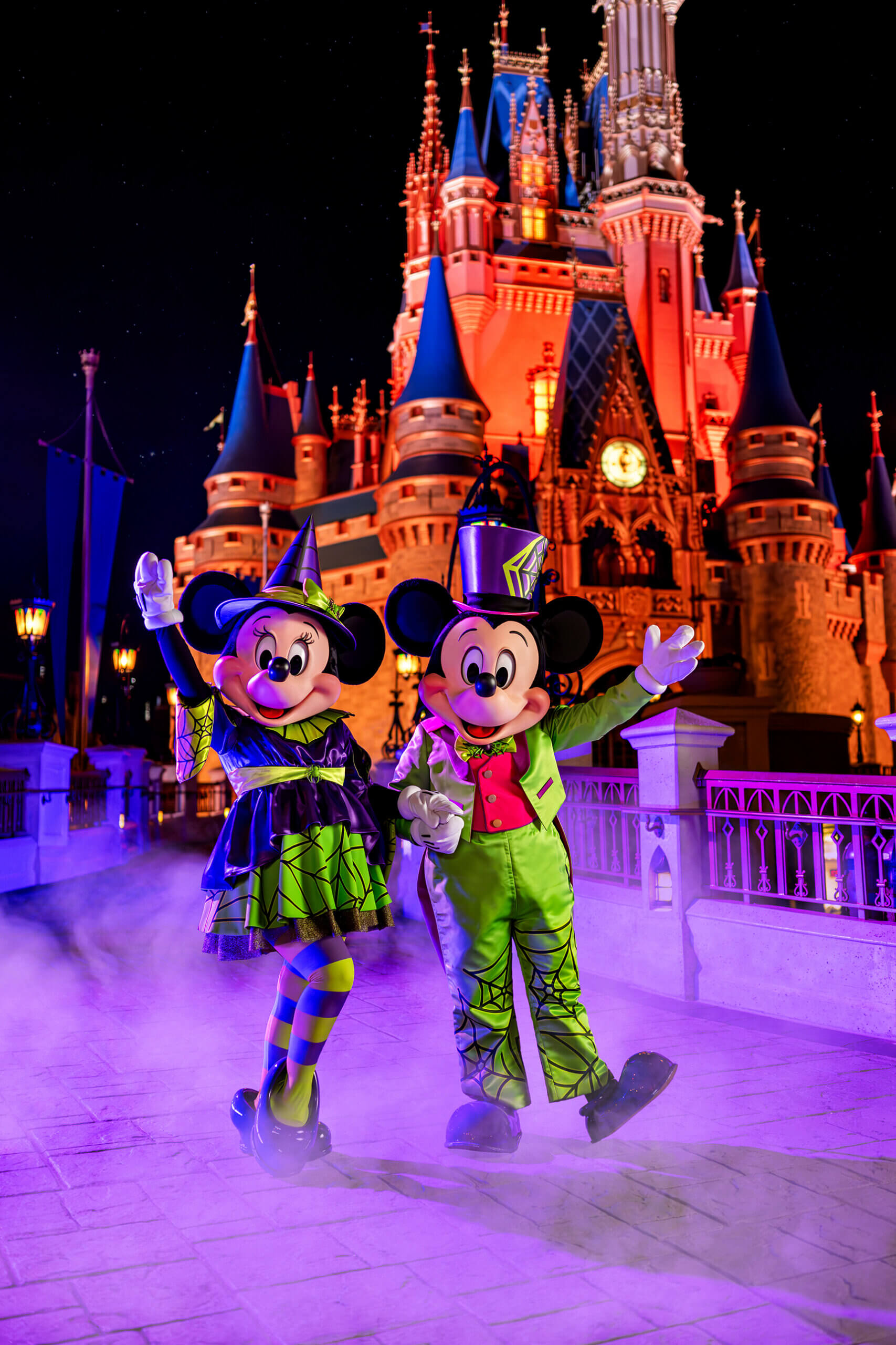Minnie and Mickey Mouse dressed in Halloween costumes stand on a misty path in front of a brightly lit castle at night, waving and smiling.