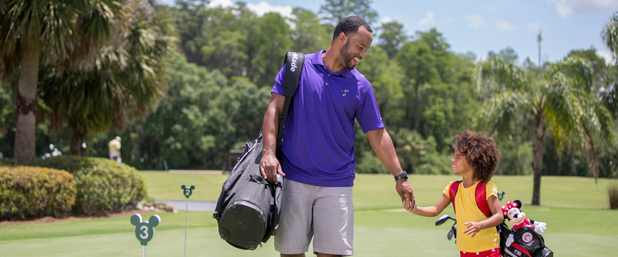 Ein Mann und ein junges Mädchen, beide mit Golfbags bepackt, gehen Hand in Hand über einen Golfplatz. An der Tasche des Mädchens ist ein Plüschtier befestigt. Sie lächeln einander an, umgeben von Bäumen und grünem Gras an einem sonnigen Tag.