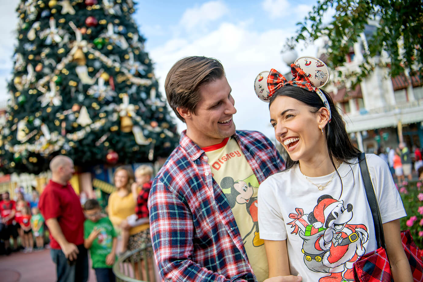 Um casal sorridente, vestindo roupas festivas com tema da Disney, está ao lado de uma grande árvore de Natal decorada ao ar livre, com outras pessoas e enfeites natalinos ao fundo.