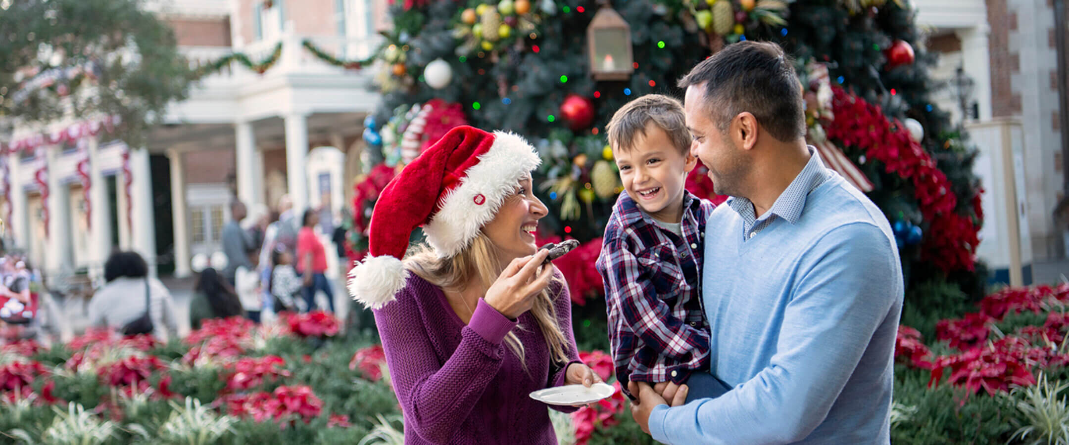 Uma família sorridente está em frente a uma grande árvore de Natal enfeitada. A mulher usa um chapéu de Papai Noel e segura um prato de sobremesa, o homem segura uma criança que ri, e decorações festivas são visíveis ao fundo.