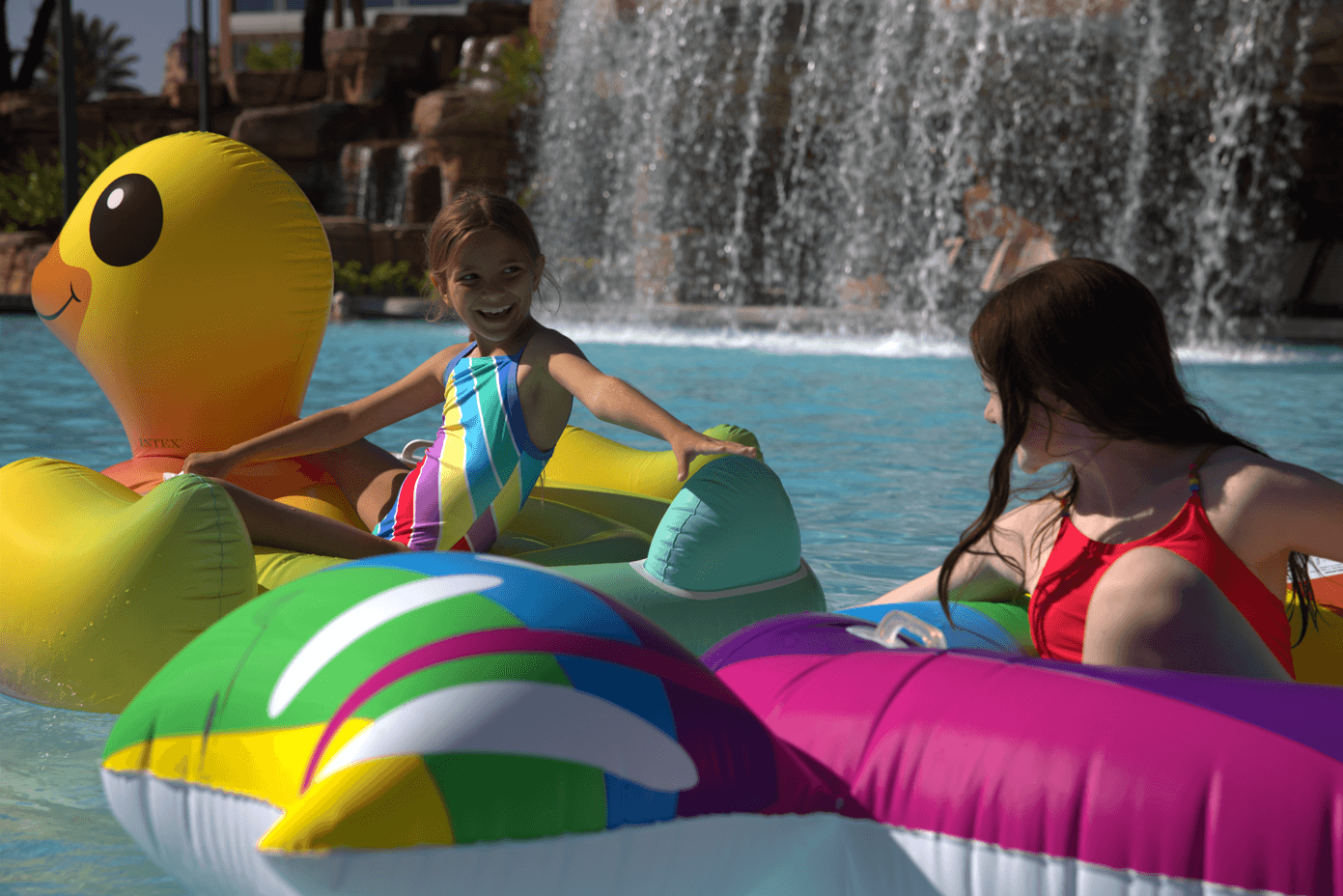 In una giornata di sole, due ragazze in costume da bagno galleggiano su colorati gonfiabili in piscina, sorridendo e giocando vicino a una cascata sullo sfondo.