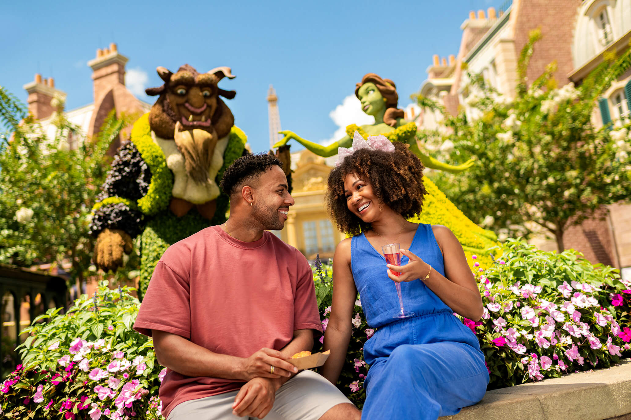 Um casal sorridente está sentado em um canteiro de flores, saboreando lanches e bebidas, com grandes figuras topiárias da Bela e a Fera ao fundo, num dia ensolarado.