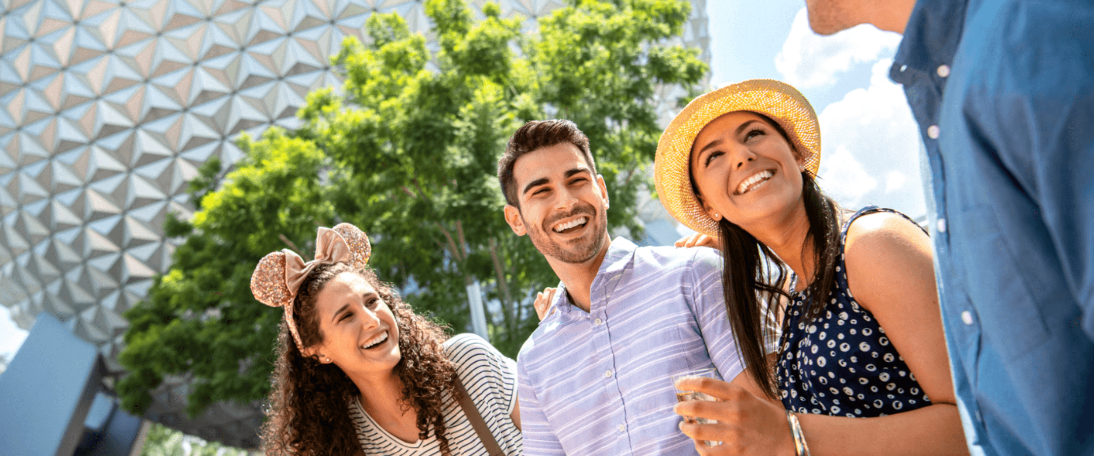 Quatro jovens estão sentados e sorriem juntos em frente a uma grande cúpula geométrica, com árvores ao fundo. Uma mulher usa orelhas de rato, outra tem um chapéu amarelo, e elas parecem estar saboreando lanches e bebidas ao ar livre.