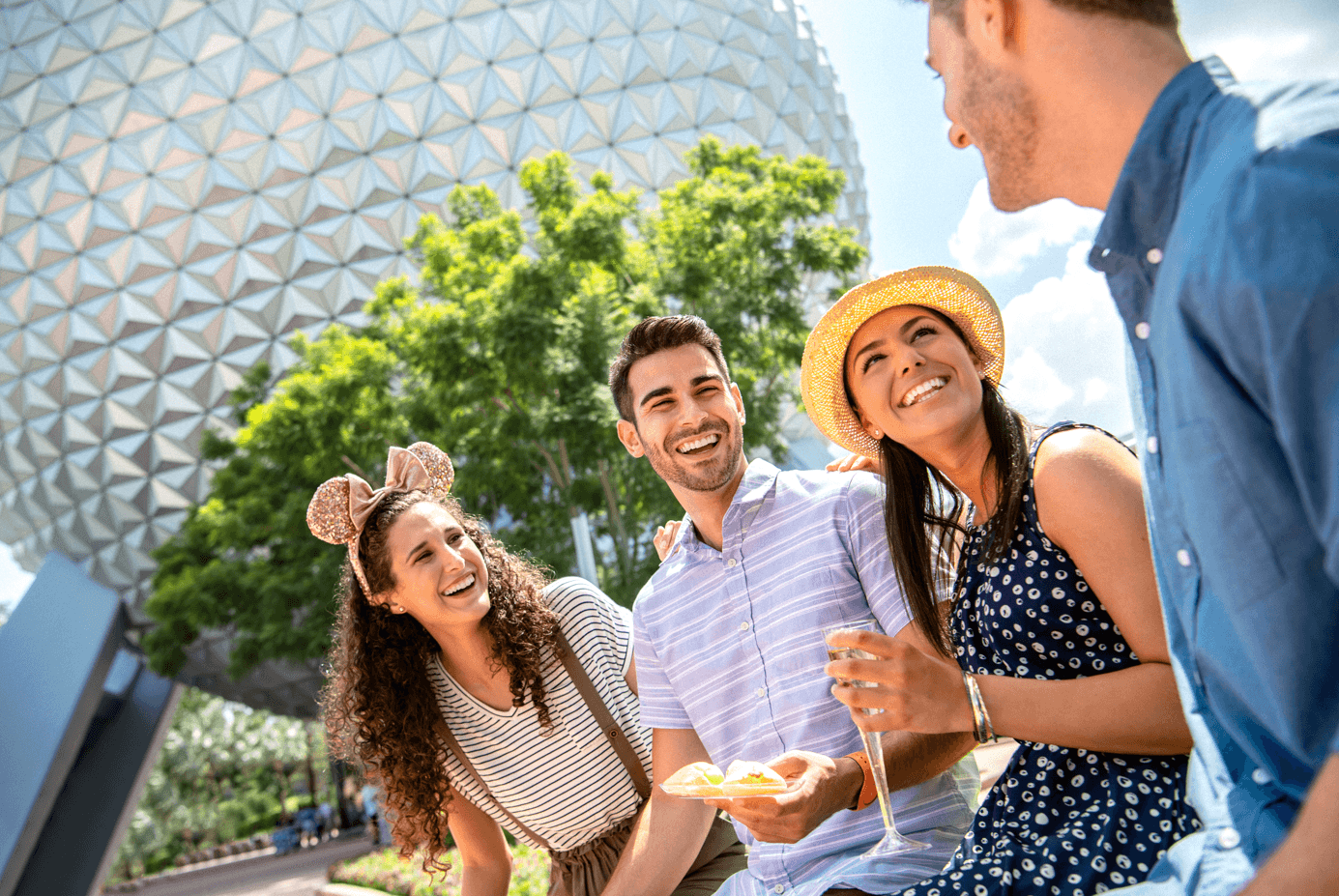 Quatro jovens estão sentados e sorriem juntos em frente a uma grande cúpula geométrica, com árvores ao fundo. Uma mulher usa orelhas de rato, outra tem um chapéu amarelo, e elas parecem estar saboreando lanches e bebidas ao ar livre.