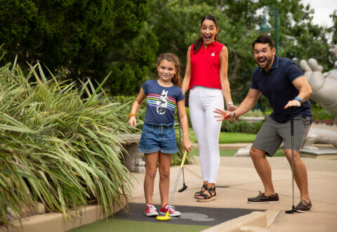 Une petite fille s'apprête à frapper la balle sur un parcours de minigolf, tandis que deux adultes derrière elle, un club à la main, sourient et réagissent avec enthousiasme. Ils sont entourés d'une végétation luxuriante par une journée ensoleillée.