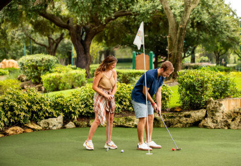 Une femme et un homme jouent au minigolf sur un parcours verdoyant, entouré d'arbres et d'arbustes luxuriants. L'homme est en train de putter tandis que la femme se tient à proximité, l'observant en souriant. On aperçoit un drapeau blanc à l'arrière-plan.