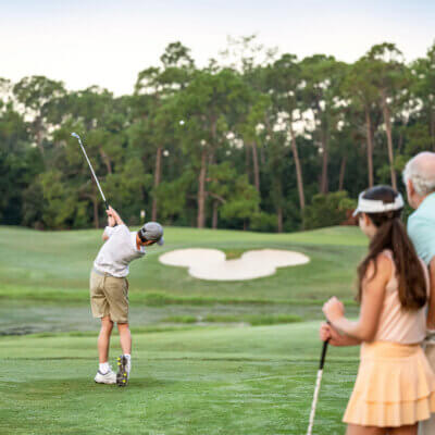 A young boy swings a golf club on a lush course while three people—an older man, a younger woman, and another man—watch him from behind. Tall trees and a sand bunker are visible in the background.