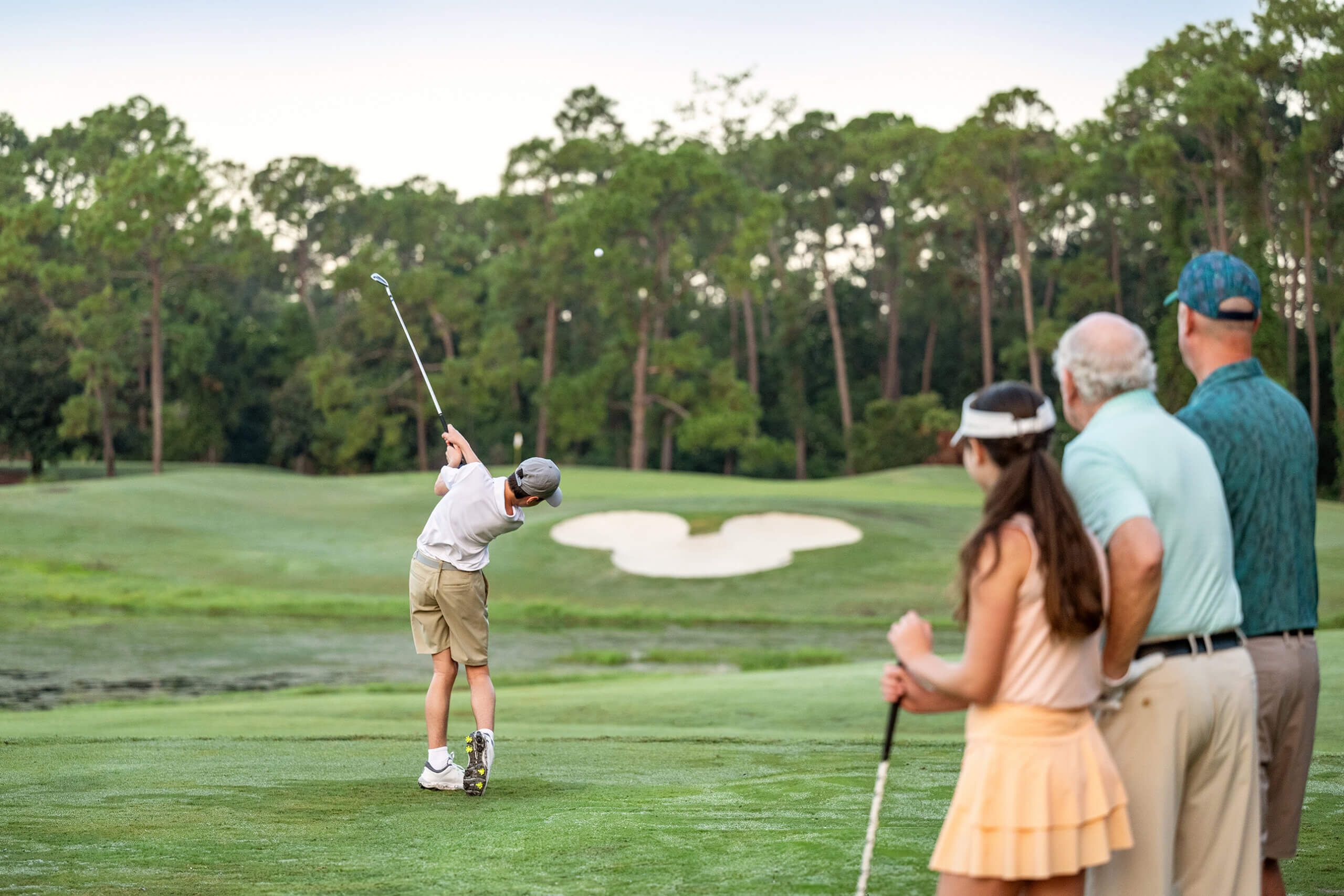 A young boy swings a golf club on a lush course while three people—an older man, a younger woman, and another man—watch him from behind. Tall trees and a sand bunker are visible in the background.