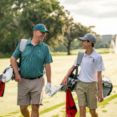 Two people, likely a father and son, walk side by side on a golf course carrying golf bags. They are smiling at each other, both dressed in shorts and polo shirts. The course is green with trees and a fountain in the background.