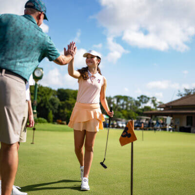 A young girl and an adult golfer high-five on a putting green under a blue sky. The girl is smiling, holding a putter, and wearing a peach skirt and white cap. A golf flag and clubhouse are visible in the background.