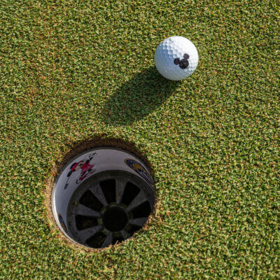 A golf ball with a Mickey Mouse logo rests on the green near a golf hole, which has stickers of Mickey Mouse on its rim. The sun casts a shadow behind the ball.