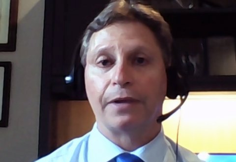 A man wearing a headset and a blue tie sits indoors, appearing to speak or listen during a video call. There is a dark piece of furniture and a lit area in the background.