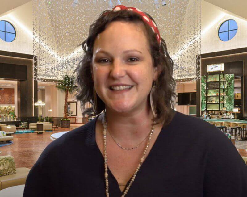 A woman with curly brown hair and a red headband smiles at the camera in a modern, well-lit lobby with large windows, hanging lights, and stylish furniture in the background.