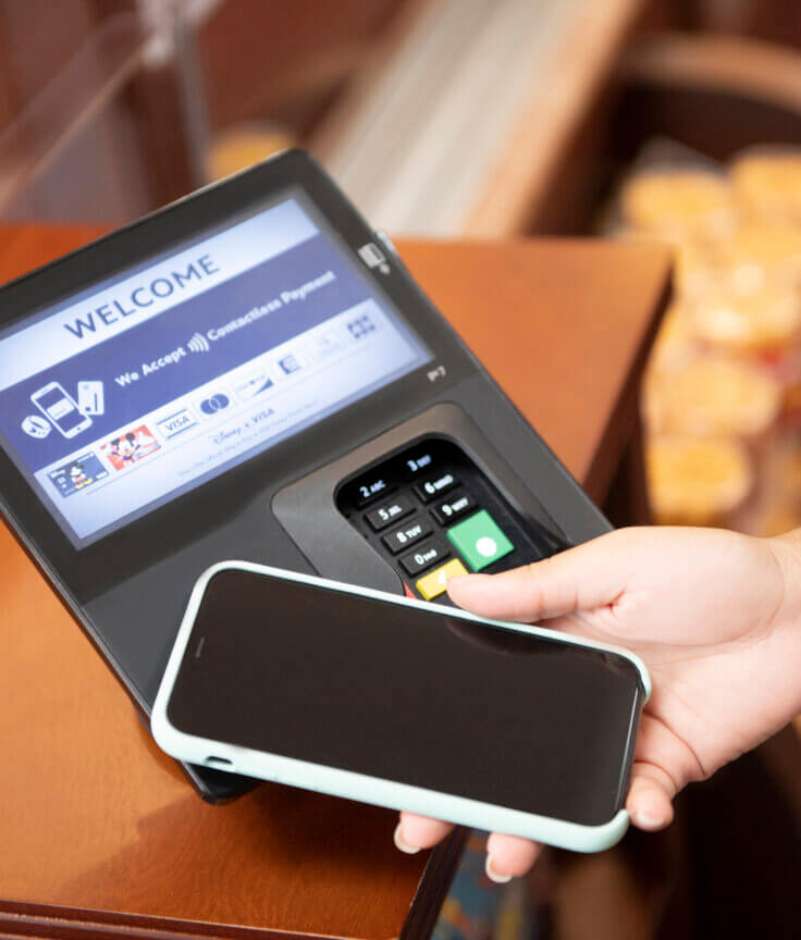 Uma pessoa aproxima um smartphone de um terminal de pagamento para efetuar um pagamento sem contato. A tela do terminal exibe uma mensagem de boas-vindas e as opções de pagamento.