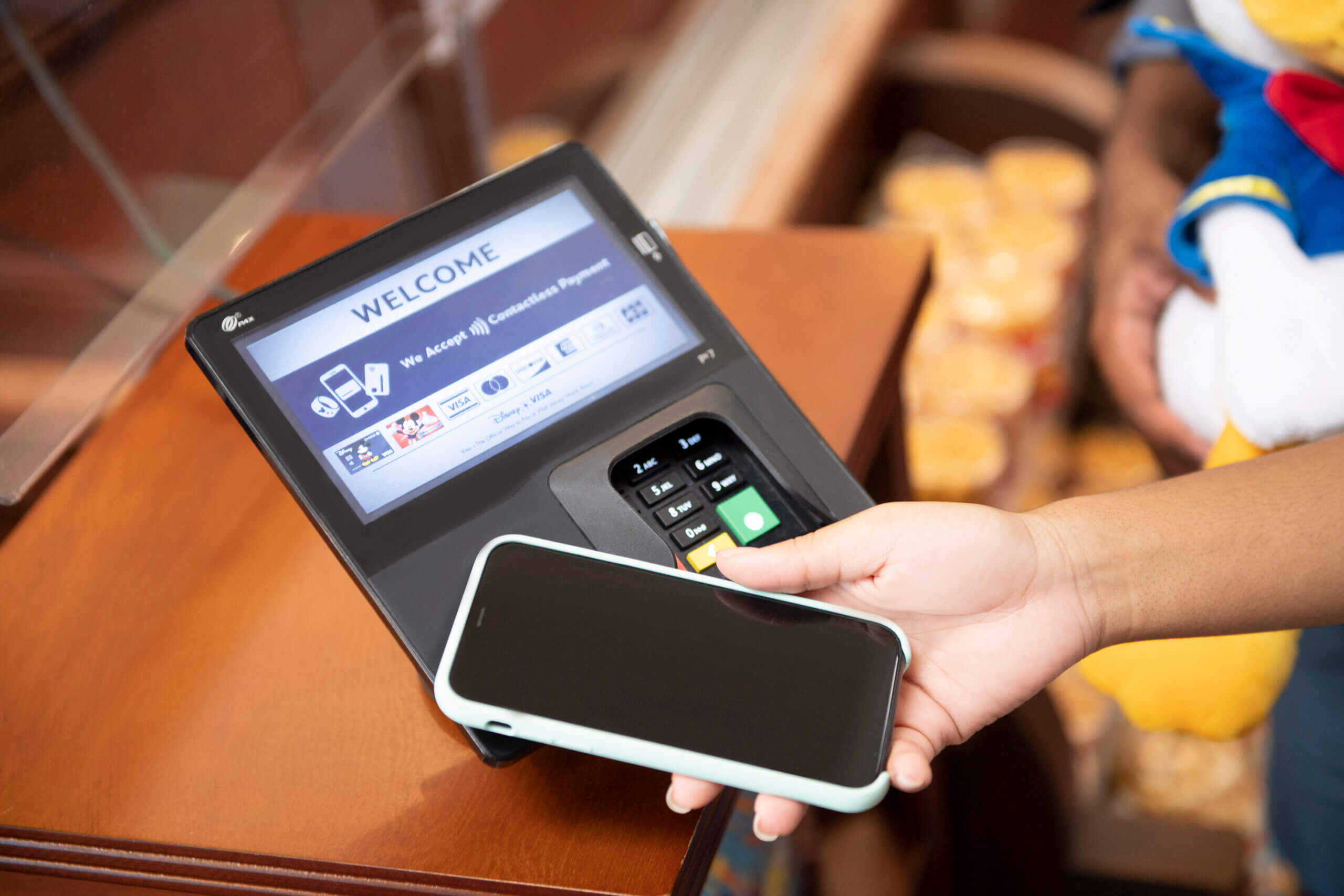 A person holds a smartphone near a payment terminal to make a contactless payment. The screen on the terminal displays a welcome message and payment options.