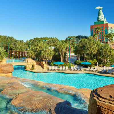 A large resort pool with rock features, surrounded by palm trees, lounge chairs, and turquoise umbrellas. In the background is a hotel building with a dolphin sculpture on the roof against a clear blue sky.