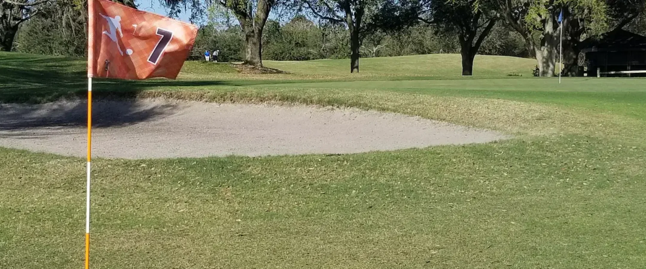 Un ballon de football jaune et noir repose près d'un trou de golf orné d'un drapeau portant le numéro 7, sur un parcours verdoyant, à côté d'un bunker de sable et entouré d'arbres.