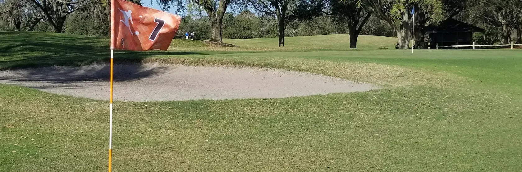 Ein gelb-schwarzer Fußball liegt in der Nähe eines Golflochs mit einer Fahne, auf der die Zahl 7 steht, auf einem grasbewachsenen Platz, neben einem Sandbunker und umgeben von Bäumen.